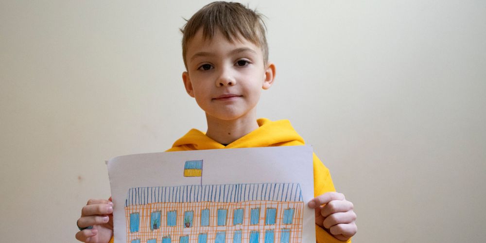 A young Ukranian boy holds up the drawing of his school. It has a blue and gold Ukranian flag flying on the roof